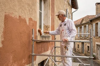 Peintre professionnel appliquant une peinture terracotta sur une façade en pierre dans un village français, debout sur un échafaudage